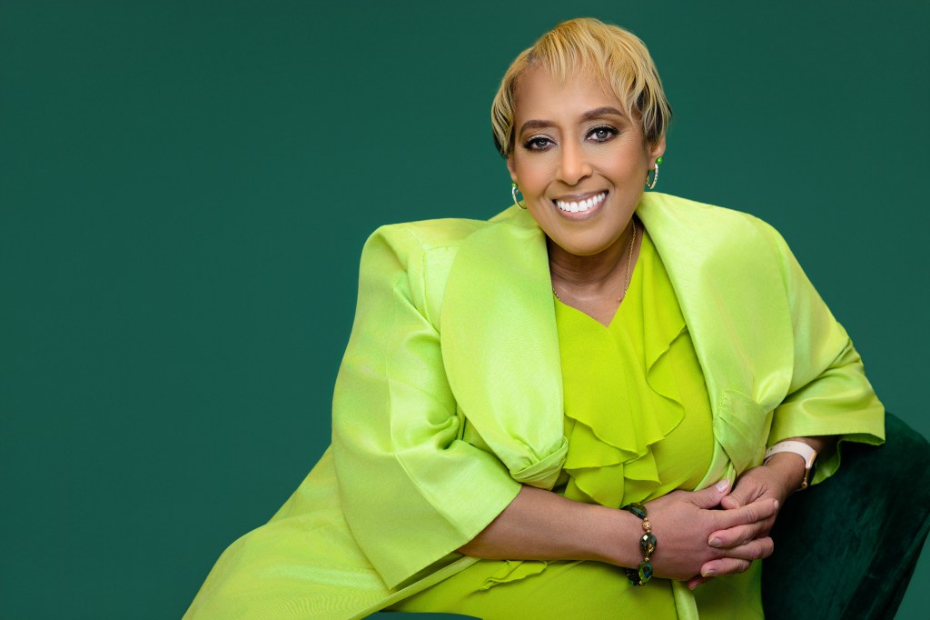 Photo of a smiling woman wearing a lime blouse and jacket, sitting sideways on a chair and leaning to her left. 